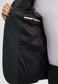 Giacca da abito verde scuro con foderato in satin nero, caratterizzata da un accento rosso sulla tasca interna, etichettata "FINEST STRETCH FABRIC."