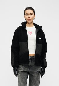 Μαύρο fleece μπουφάν με ψηλό γιακά, με τμήμα από λείο μαύρο ύφασμα, φορεμένο πάνω από λευκό πουκάμισο με ροζ καρδιά. Μαύρα γάντια.