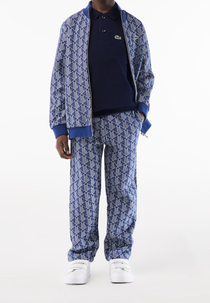 Personne portant une veste et un pantalon à motif géométrique bleu, un polo bleu marine avec un logo et des baskets blanches, debout les mains détendues.