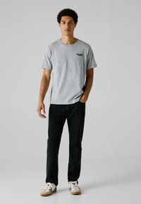 Camiseta de manga corta gris con el logo negro de "RED BULL", combinada con jeans oscuros y zapatillas blancas con detalles en negro y suela de goma.