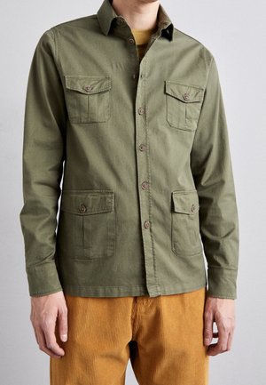 Mand iført olivengrøn knap-up overshirt med fire knaplommer og sennepsfarvede fløjlsbukser mod ensfarvet baggrund.
