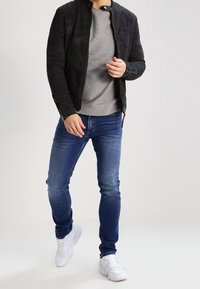 Chaqueta acolchada negra con un cuello alto, camiseta de rayas gris debajo, jeans ajustados azules y zapatillas blancas. El conjunto presenta un estilo casual.