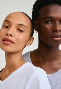 Due persone molto vicine, entrambe indossano abbigliamento semplice; una in una maglietta bianca, l'altra in una maglietta grigia. Orecchini visibili.