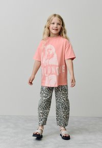 Nuori tyttö, jolla on pitkät vaaleat hiukset, on pukeutunut vaaleanpunaisiin Blondie-grafiikkapaitaan, leopardikuvioisiin housuihin ja valkoisiin sandaaleihin, seisten harmaata taustaa vasten.