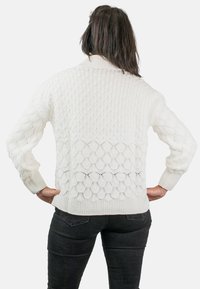 Persona con cabello largo y oscuro, vistiendo un suéter blanco de punto texturizado y jeans negros, de pie de espaldas con las manos en las caderas.