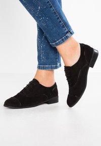 Scarpe oxford in suede nero con una finitura liscia, punta arrotondata, tacco basso e design con lacci, caratterizzate da un dettaglio a cappuccio in contrasto.