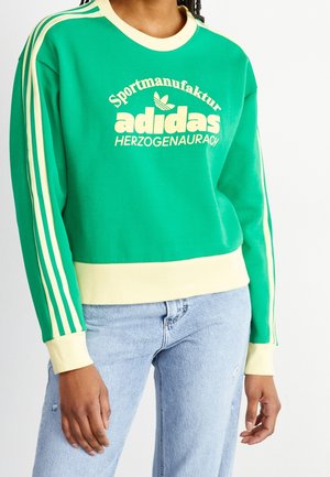 Persoon met een groene Adidas-trui met crèmekleurige strepen op de mouwen en een crème zoom, gecombineerd met lichtblauwe spijkerbroek.