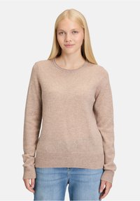 Beżowy sweter z okrągłym dekoltem, wykonany z miękkiego materiału, z długimi rękawami oraz ściągaczami przy mankietach i u dołu. Połączony z jasnoniebieskimi dżinsami.