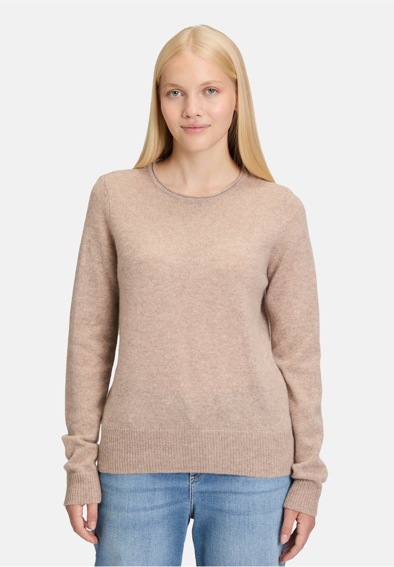 Beżowy sweter z okrągłym dekoltem, wykonany z miękkiego materiału, z długimi rękawami oraz ściągaczami przy mankietach i u dołu. Połączony z jasnoniebieskimi dżinsami.