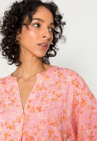 Blusa floral rosa com uma textura macia, apresentando padrões de flores laranja e brancos, um decote em V e mangas curtas.