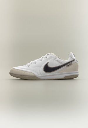 Chaussure de football indoor Nike Tiempo blanche basse avec logo Swoosh noir et détails en daim beige à l’avant et au talon.