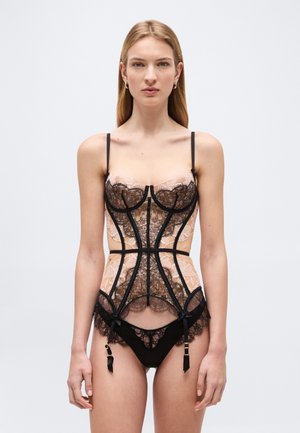 Kobieta ubrana w przejrzysty, beżowo-czarny koronkowy gorset z podwiązkami oraz pasujące koronkowe majtki, stojąca na jednolitym jasnym tle.