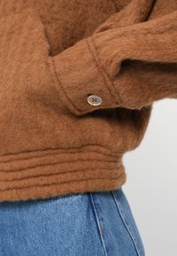 Brun, fluffy sweater med ribbet kant. Har en rund krave og en knapdetalje på ærmet, parret med blå denim.