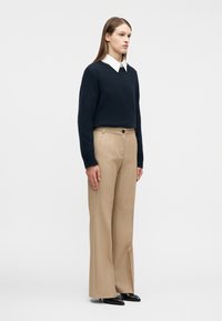 Γυναίκα που στέκεται, φοράει μπλε πουλόβερ πάνω από λευκό πουκάμισο με γιακά, μπεζ παντελόνι με φαρδιά μπατζάκια και μαύρα μυτερά παπούτσια σε απλό φόντο.