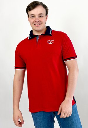 Jeune homme souriant, portant un polo rouge avec le logo "Black Wellis" et un jean bleu, debout devant un fond blanc uni.