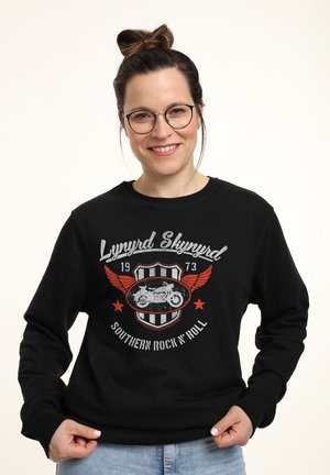 Sweat-shirt noir avec un imprimé graphique d'une moto avec des ailes, le texte "Lynyrd Skynyrd" et des éléments de style vintage. Manches longues.