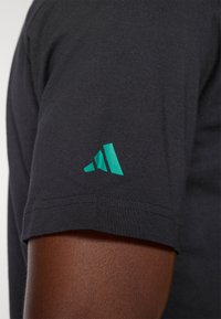 Czarny t-shirt z krótkim rękawem wykonany z miękkiej tkaniny, z turkusowym logo Adidas na lewym rękawie. Brzeg rękawa jest przeszyty dla trwałości.