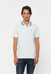 Mann i lys blå polo skjorte med svartstripet krage og mørke jeans, stående mot hvit bakgrunn.