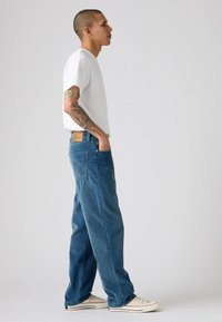 Jeans din denim albastru cu o croială lejeră, având un material texturat, design cu cinci buzunare și o etichetă din piele bej pe partea din spate a taliei. Tricou alb și adidași.