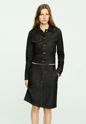 Mujer con chaqueta vaquera negra con botones y falda a juego hasta la rodilla, de pie con una mano en el bolsillo frente a un fondo simple.