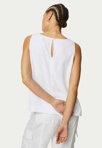 Top sans manches en lin blanc présentant une fermeture arrière en forme de trou de serrure et un ourlet droit. Le tissu a une apparence douce et texturée.