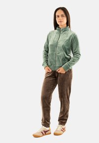 Giacca verde in velluto con zip e due tasche frontali, abbinata a pantaloni in velluto marroni e sneakers beige con righe rosse. Tessuto liscio.