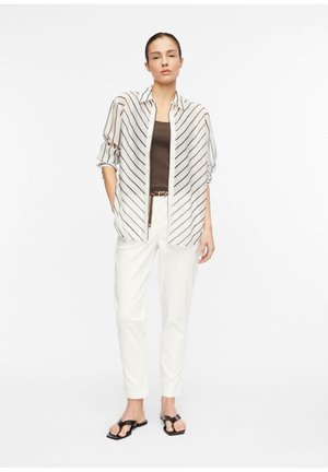 Kobieta stojąca, ubrana w białą koszulę w paski z guzikami na brązowym topie, białe spodnie z paskiem oraz czarne sandały, na jednolitym tle.