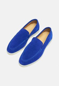 Plave nubuk slip-on cipele s bijelim gumenim potplatom, s vidljivim šavovima duž rubova i glatkom unutarnjom podstavom.