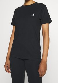 Sort bomulds T-shirt med korte ærmer, rund hals og et lille hvidt kænguru-logo på brystet. Glat tekstur og afslappet pasform.
