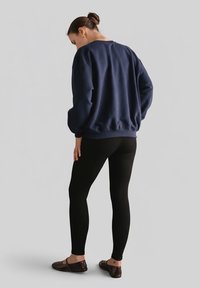 Granatowa bluza z prążkowanymi mankietami i dołem, w zestawieniu z czarnymi legginsami i ciemnobrązowymi płaskimi butami, widziana od tyłu na szarym tle.