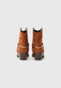 Bottines en suède marron avec un petit talon carré, ornées d'une chaîne dorée autour de la cheville et d'un design légèrement pointu.