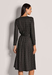 Frau mit langen dunklen Haaren, die ein schwarzes Midikleid mit beigem kreisförmigem Kettenmuster und langen Ärmeln trägt, steht vor einer schlichten hellen Wand.