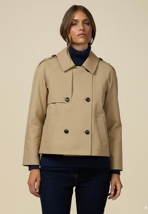 Chaqueta beige de doble botonadura con grandes botones negros, con cuello y hombreras. Lleva puesta una sudadera de canalé azul marino, combinada con jeans oscuros.