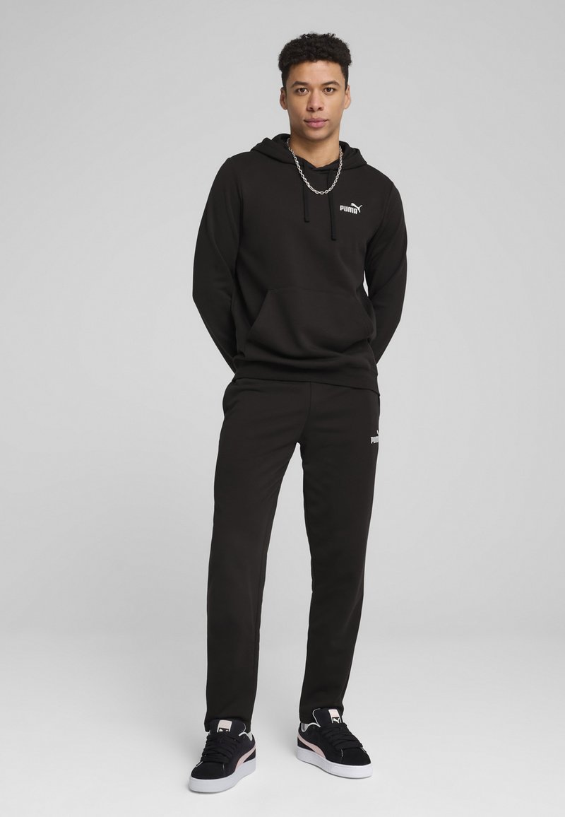 Um hoodie preto e umas calças de treino com um design simples; possui um bolso frontal e um logo no peito; combinado com ténis pretos e brancos.