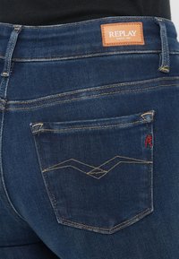 Jeans de mezclilla azul oscuro con una etiqueta de cuero marrón que dice "REPLAY". Presentan un bolsillo trasero con costuras amarillas y un detalle bordado rojo.