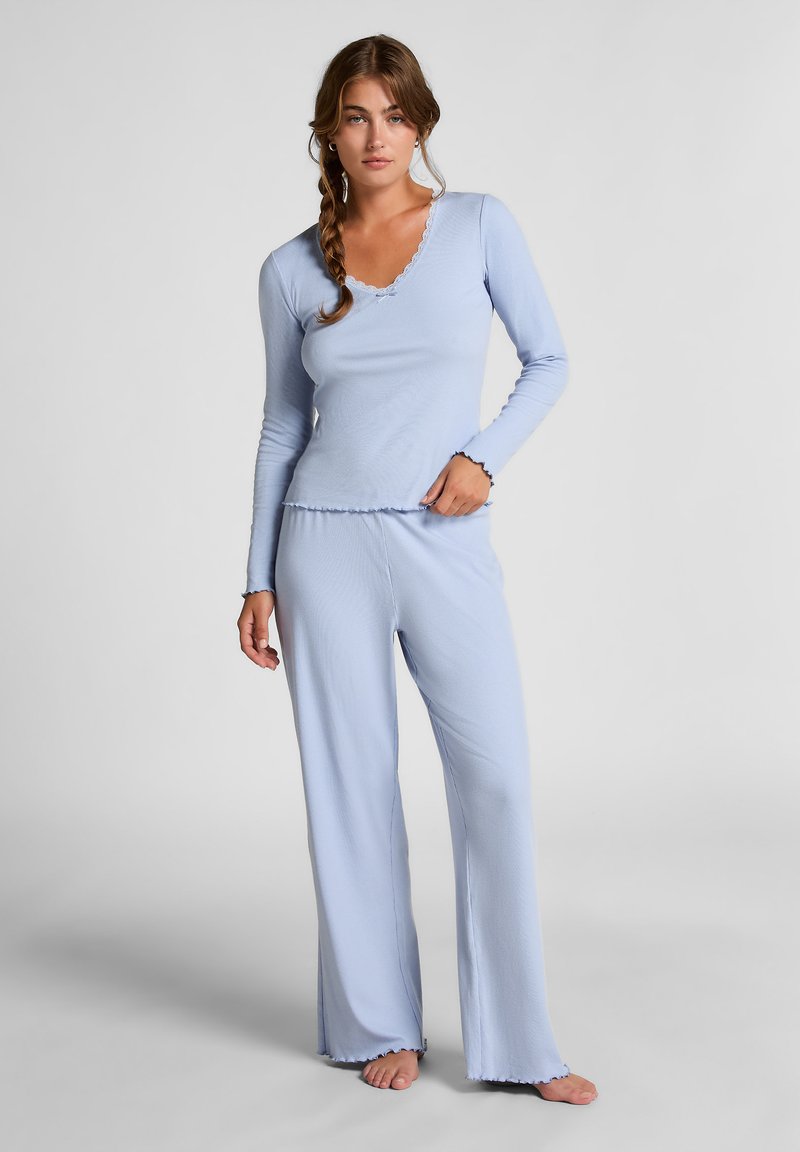 Vrouw met gevlochten haar, gekleed in een lichtblauwe lange mouw top en brede broek, die blootsvoets tegen een effen achtergrond staat.