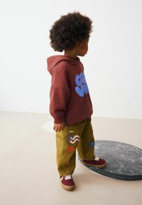 Enfant portant un sweat à capuche bordeaux avec des lettres bleues, un pantalon vert olive avec des motifs colorés et des baskets bordeaux avec des accents blancs.