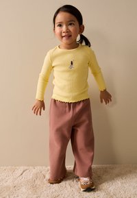 Žuta duga majica s rebrastim uzorkom i valovitim rubovima s grafikom psa, u kombinaciji s širokim ružičastim trenirkama i tenisicama s uzorkom.