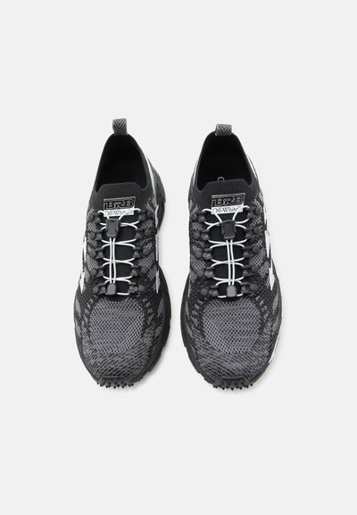 Ζευγάρι μαύρα και γκρι πλεκτά αθλητικά παπούτσια με λευκά ελαστικά κορδόνια και ετικέτες με λογότυπο, από πάνω σε λευκό φόντο.