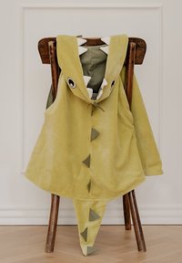 Capa con capucha de temática de dinosaurio verde, con acentos de dientes blancos, picos triangulares y textura de felpa, exhibida en una silla de madera.