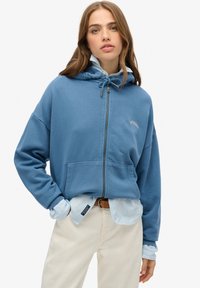 Felpa blu con zip, dotata di coulisse, tasche frontali e dettaglio del logo. Indossata sopra una camicia a righe blu chiaro e abbinata a pantaloni color crema.