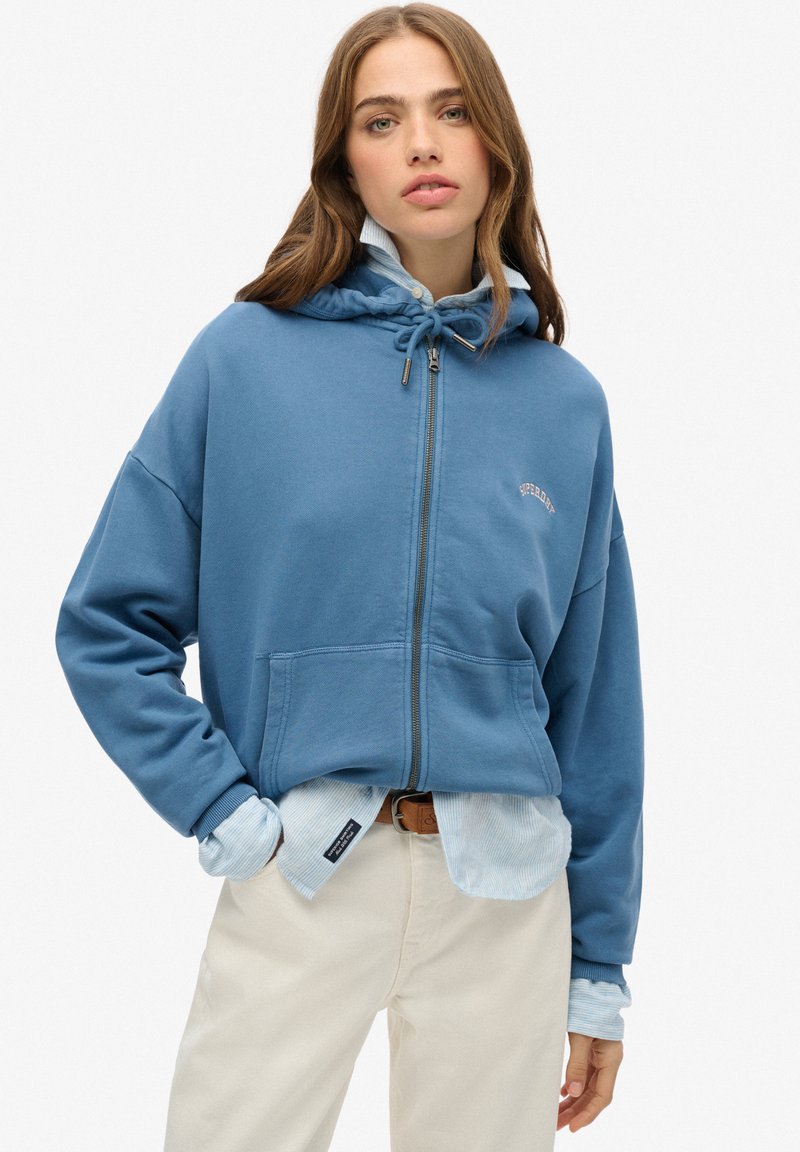 Felpa blu con zip, dotata di coulisse, tasche frontali e dettaglio del logo. Indossata sopra una camicia a righe blu chiaro e abbinata a pantaloni color crema.