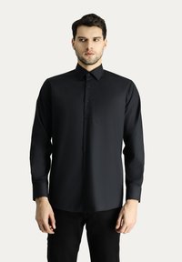Camicia nera a maniche lunghe con bottoni, colletto a punta, taschino frontale e tessuto liscio e strutturato. Il modello indossa dei pantaloni neri.