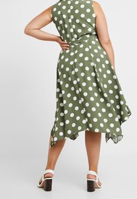 Grünes gepunktetes Kleid mit asymmetrischem, fließendem Saum und taillierter Taille. Verfügt über ein ärmelloses Design und eine glatte Stoffoberfläche.