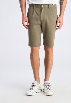 Uomo che indossa pantaloncini su misura color verde oliva fino al ginocchio e scarpe da ginnastica bianche con dettagli neri e blu.