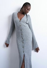 Femme aux cheveux tressés portant une longue robe en tricot gris côtelée boutonnée, debout contre un mur blanc, regardant vers le bas.