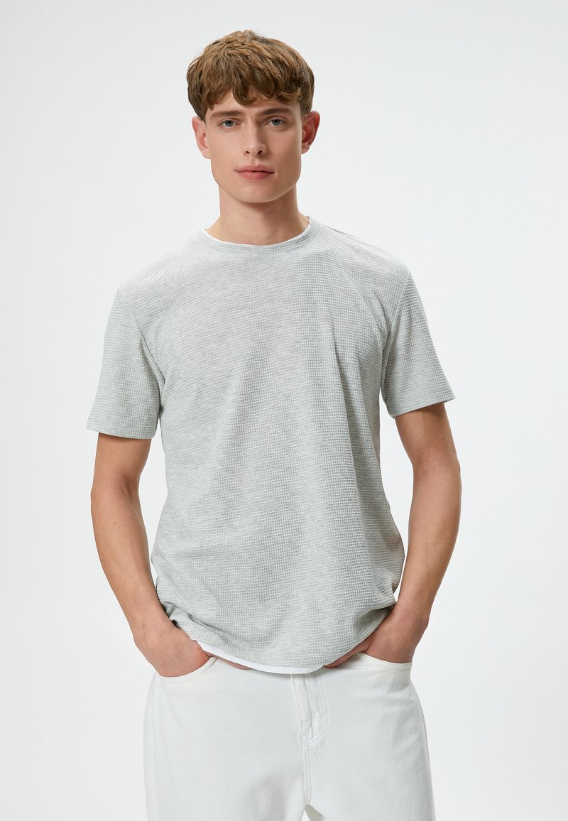 Grijze, textuurgegeven t-shirt met korte mouwen en een ronde halslijn, met een iets losse pasvorm, gecombineerd met witte broek, weergegeven tegen een effen achtergrond.