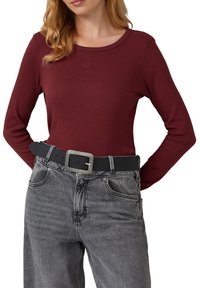Haut long à manches rouges avec une texture côtelée, un col rond et un détail de logo. Associé à un jean gris taille haute et une ceinture noire avec une boucle en métal argenté.