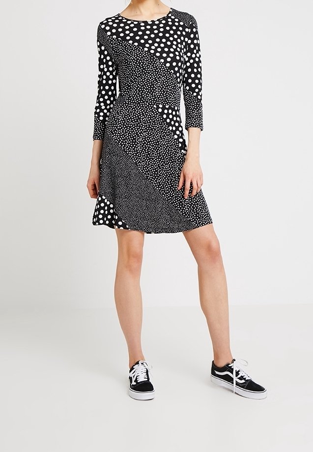 Femme portant une robe à pois noirs et blancs avec des manches trois-quarts et des baskets noires avec des lacets blancs.