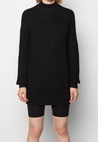 Pull en tricot noir avec un col montant, texture côtelée et manches longues, associé à un short de cyclisme noir.